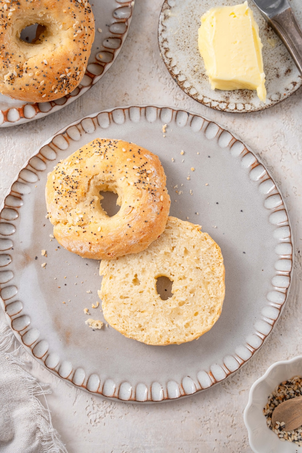 A ceramic plate with a sliced cottage cheese bagel on a marble counter. There is another ceramic plate with butter and whole bagels on the side.