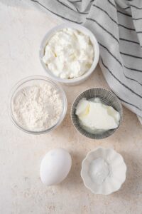 An overhead shot of several bowls of various sizes containing ingredients for cottage cheese waffles including cottage cheese, flour, greek yogurt, and an egg.