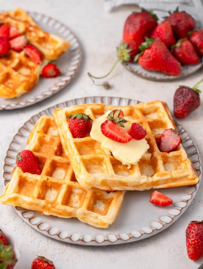 Two cottage cheese waffles on a ceramic plate topped with butter and sliced strawberries. There is another plate of waffles and a plate of strawberries in the background.