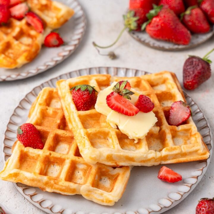 Two cottage cheese waffles on a ceramic plate topped with butter and sliced strawberries. There is another plate of waffles and a plate of strawberries in the background.