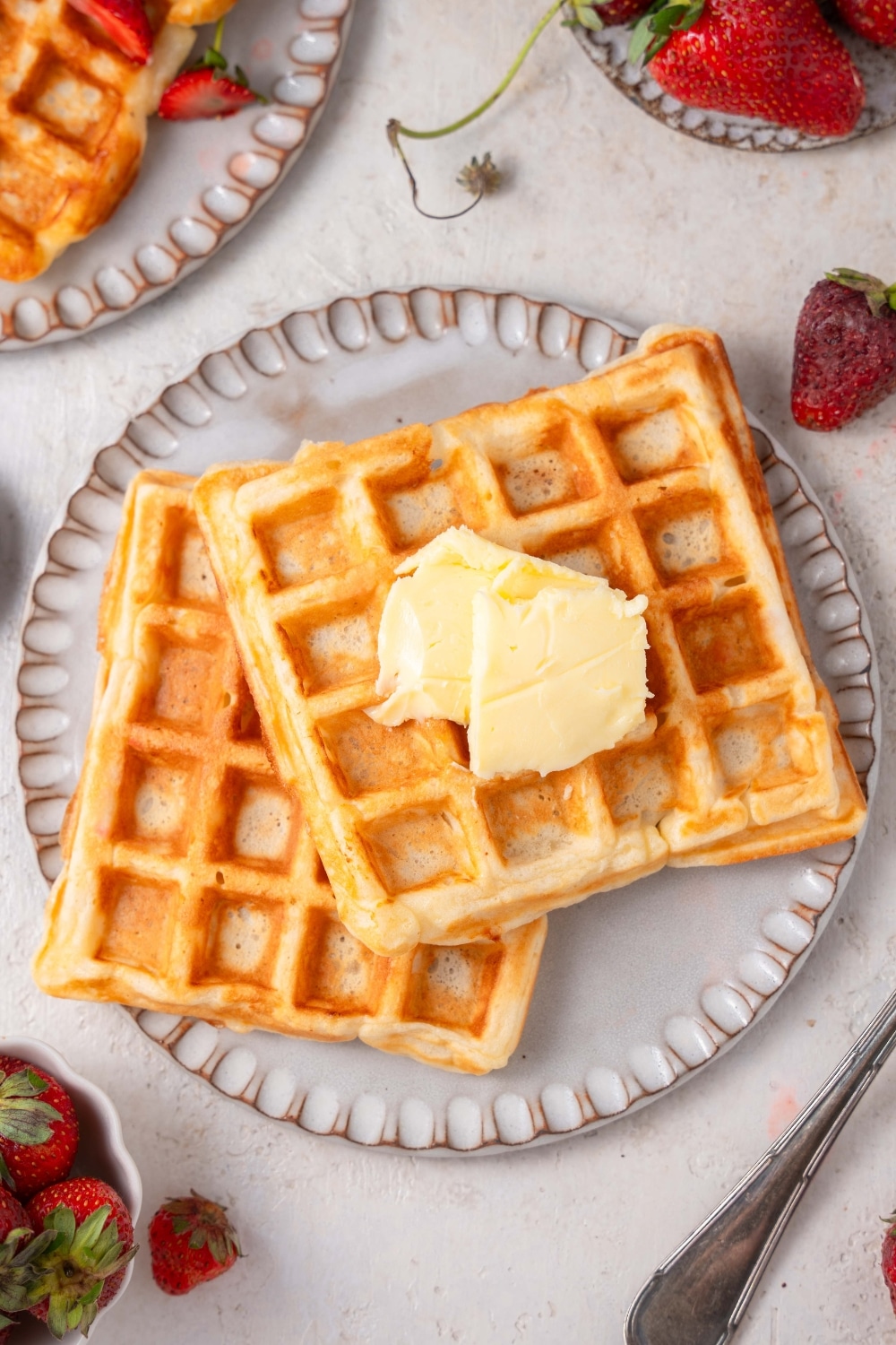 Two cottage cheese waffles on a ceramic plate topped with butter. The plate is on a marble counter with another plate of waffles and a plate of strawberries off to the side.