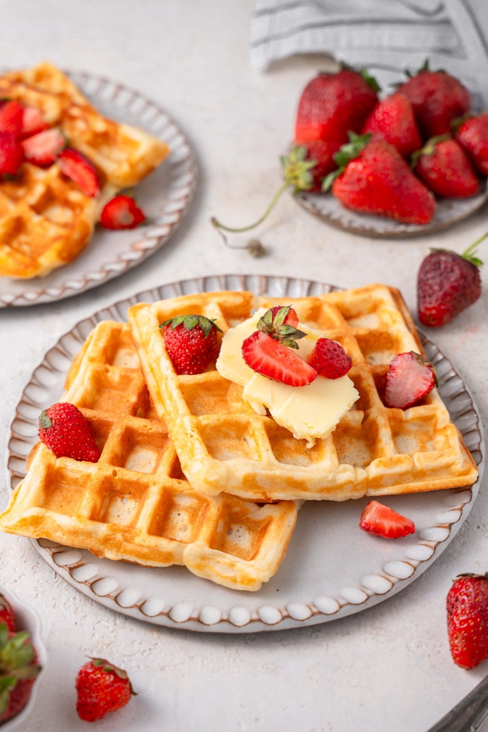 Two cottage cheese waffles on a ceramic plate topped with butter and sliced strawberries. There is another plate of waffles and a plate of strawberries in the background.