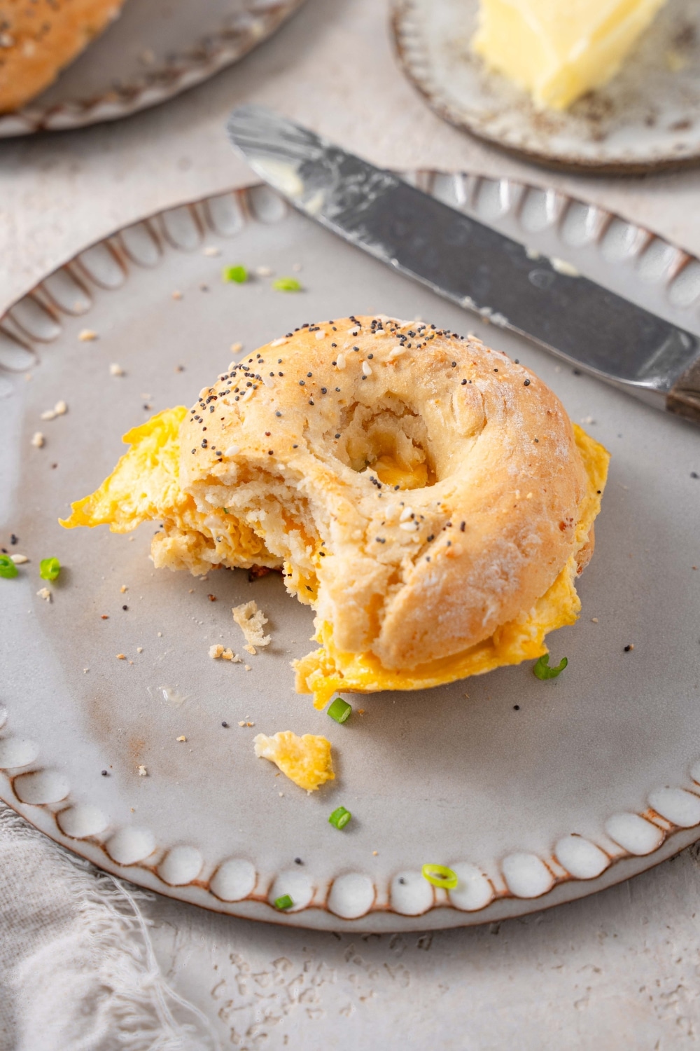 A ceramic plate with a scrambled egg breakfast sandwich using a cottage cheese bagel with everything bagel seasoning. There is a bite taken out of the sandwich with a knife on the side.