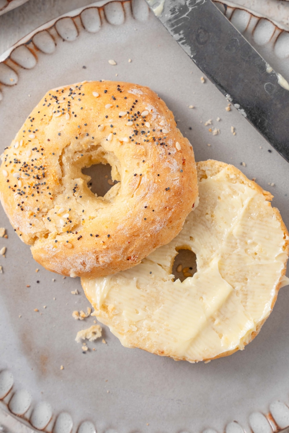 A ceramic plate with a sliced cottage cheese bagel with everything bagel seasoning and spread with butter. There is a knife on the side.