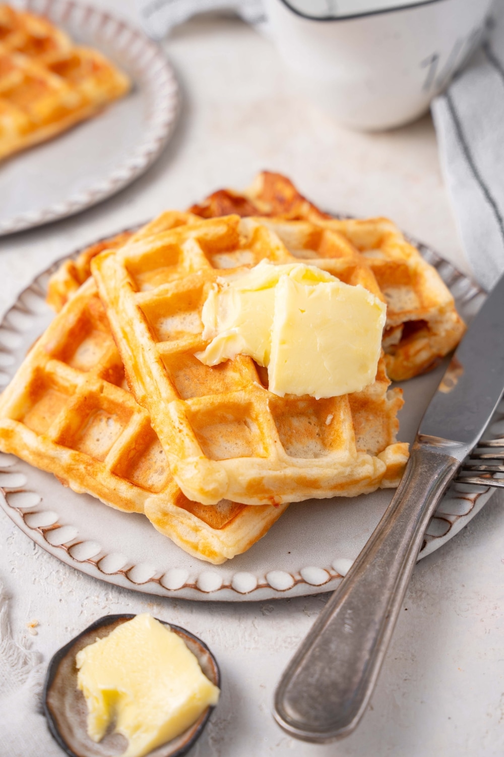 A stack of three cottage cheese waffles on a ceramic plate topped with butter. There is a knife on the plate and a bite is taken out of the waffles.