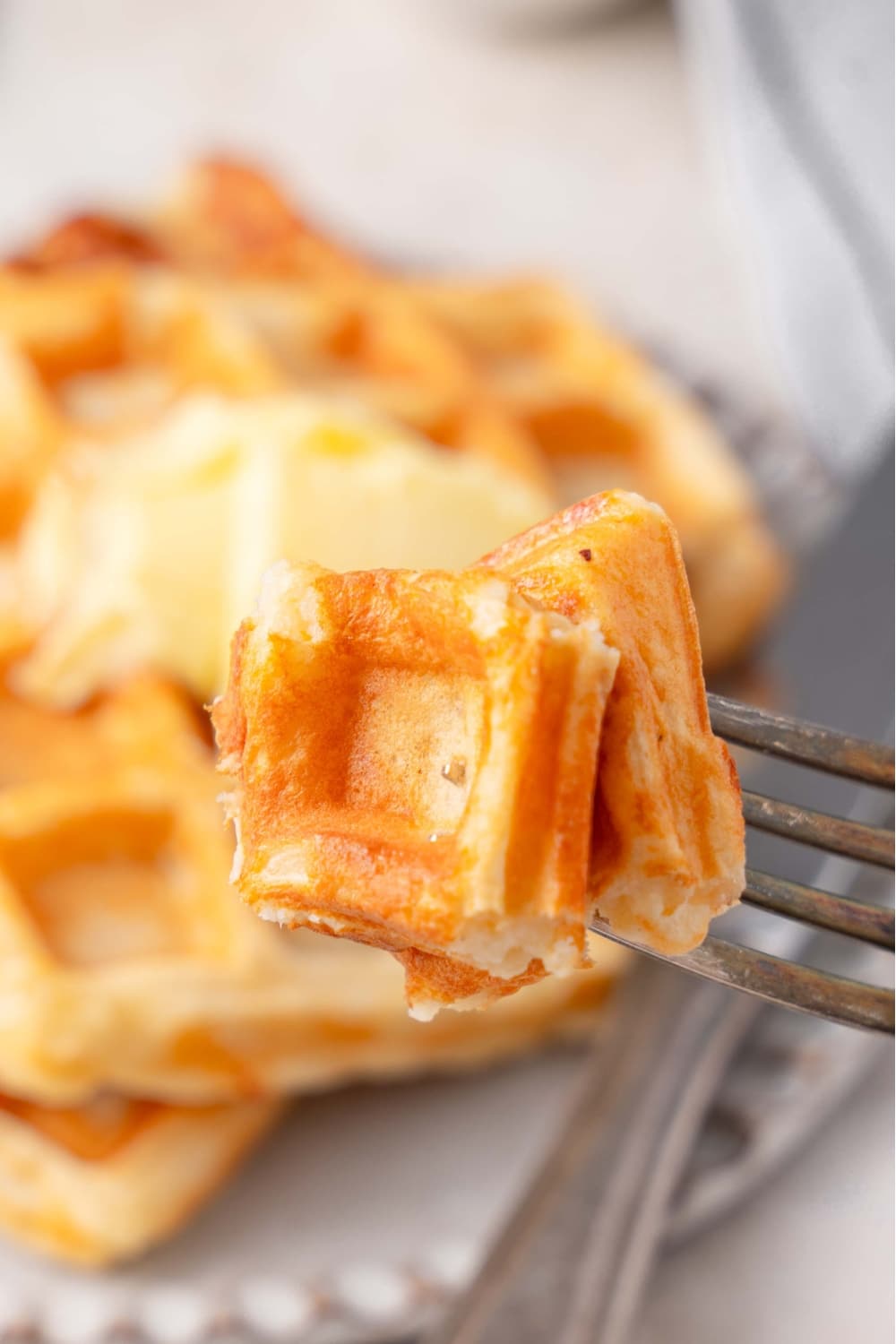 A close up of a fork with a bite of cottage cheese waffle. There is a ceramic plate with cottage cheese waffles blurred in the background.