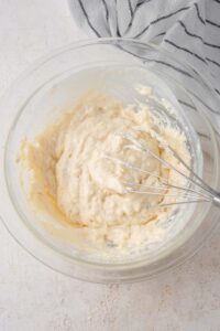 A glass bowl on a white counter with a whisk mixing cottage cheese waffle batter. There is a striped gray napkin off to the side.