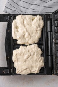 An open waffle iron with two raw cottage cheese waffles. There is a striped gray napkin off to the side.