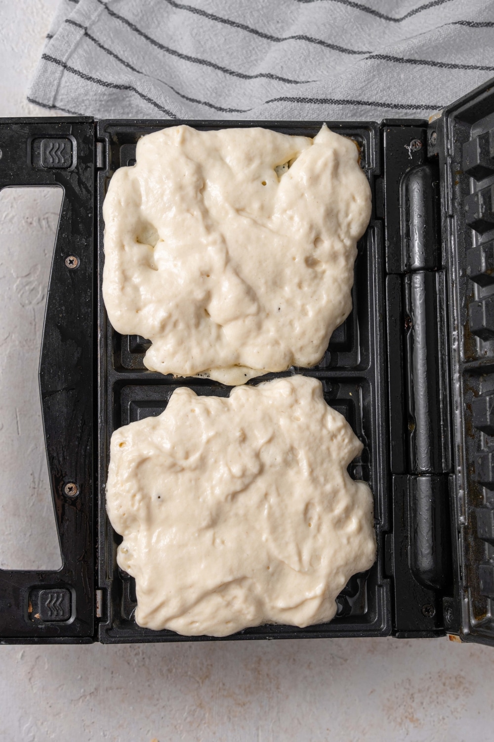 An open waffle iron with two raw cottage cheese waffles. There is a striped gray napkin off to the side.