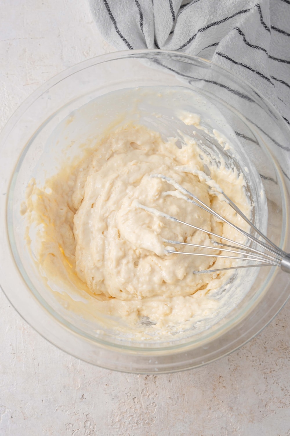A glass bowl on a white counter with a whisk mixing cottage cheese waffle batter. There is a striped gray napkin off to the side.