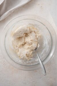 An overhead shot of a glass bowl with cottage cheese bagel dough and a spoon stirring the dough.