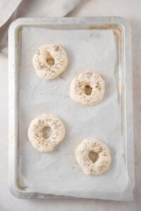 An overhead shot of a baking sheet lined with parchment paper. There are four raw cottage cheese bagels seasoned with everything bagel seasoning on the baking sheet.