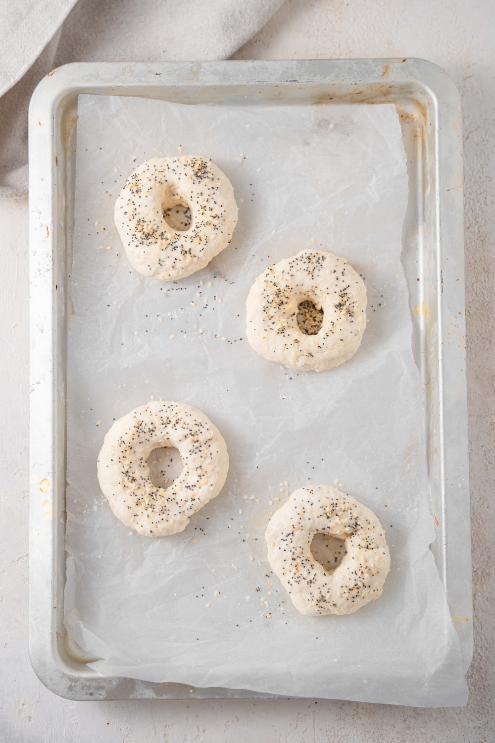 An overhead shot of a baking sheet lined with parchment paper. There are four raw cottage cheese bagels seasoned with everything bagel seasoning on the baking sheet.