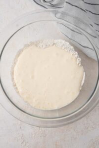 A glass bowl on a white counter filled with flour and blended cottage cheese mixture. There is a striped gray napkin off to the side.