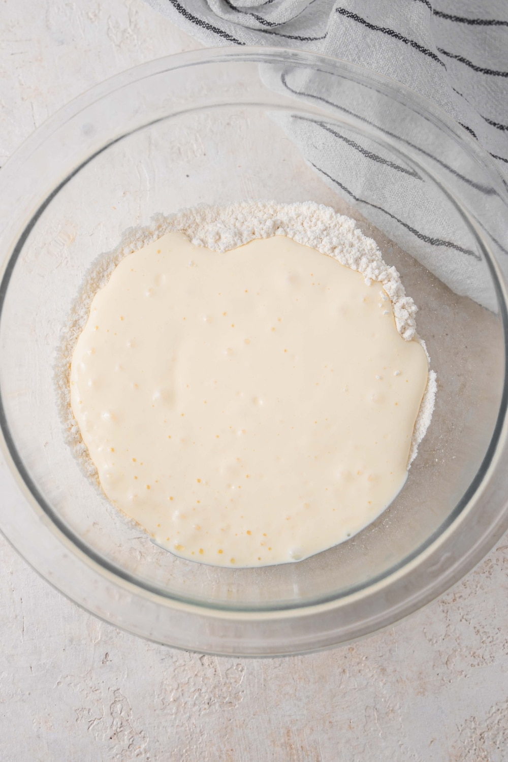 A glass bowl on a white counter filled with flour and blended cottage cheese mixture. There is a striped gray napkin off to the side.