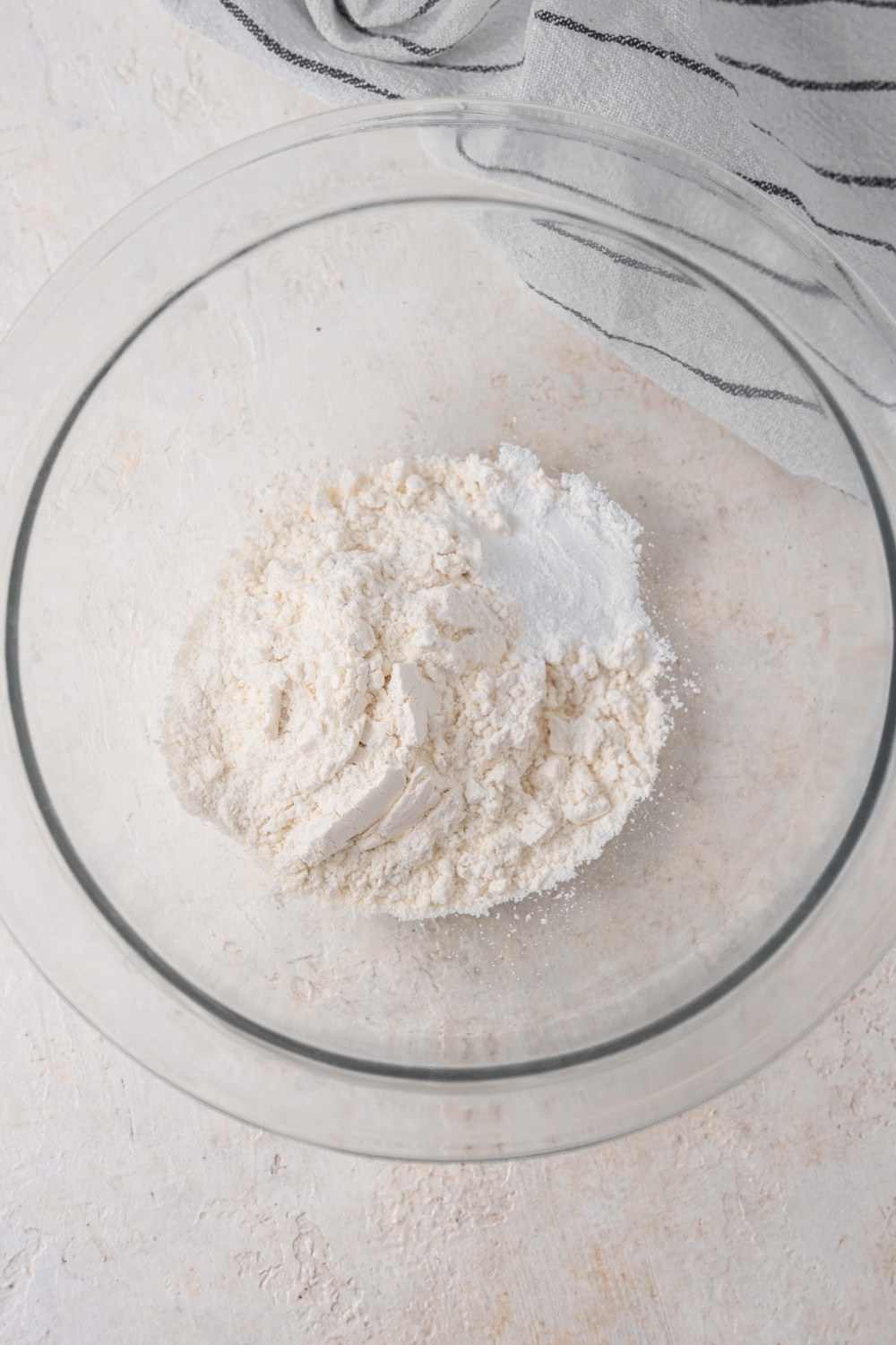A glass bowl on a white counter filled with flour and baking powder. There is a striped gray napkin off to the side.