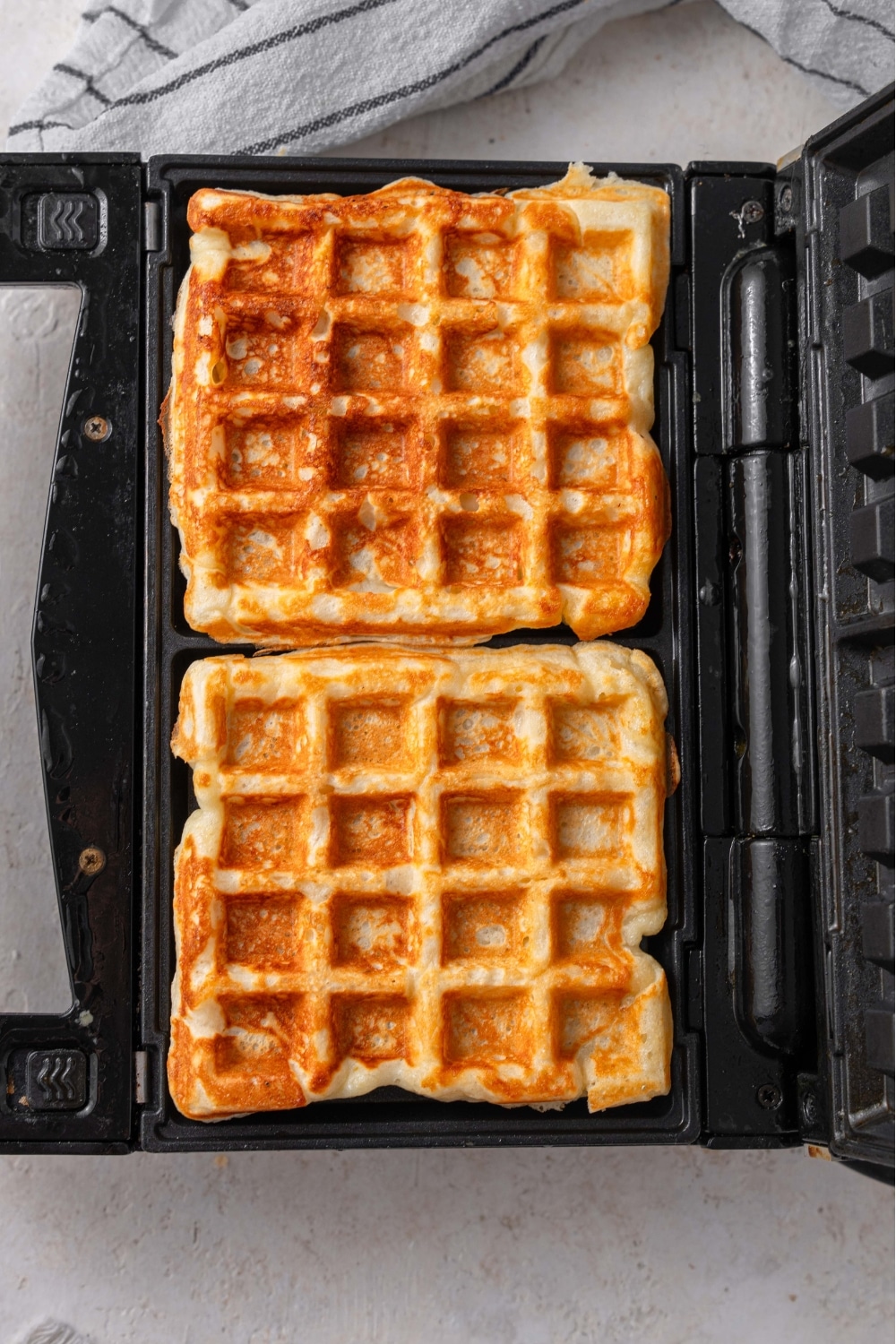 An open waffle iron with two cooked cottage cheese waffles on a white counter. There is a striped gray napkin off to the side.