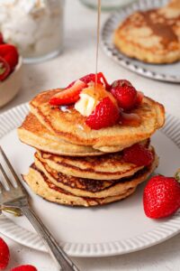 Syrup being poured over a stack of cottage cheese pancakes topped with butter and strawberries.