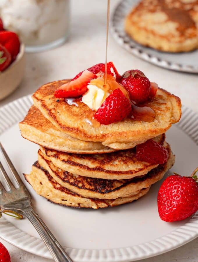 Syrup being poured over a stack of cottage cheese pancakes topped with butter and strawberries.