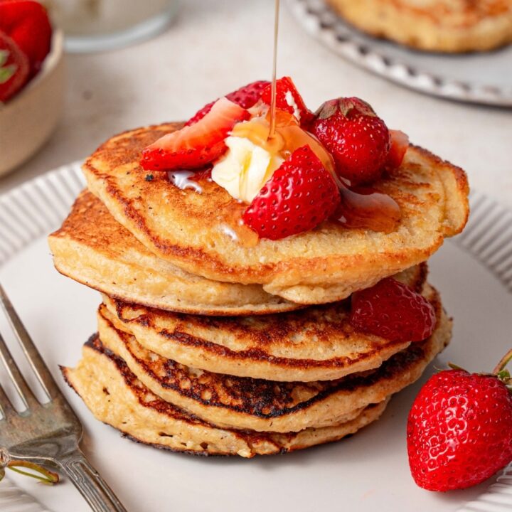 Syrup being poured over a stack of cottage cheese pancakes topped with butter and strawberries.