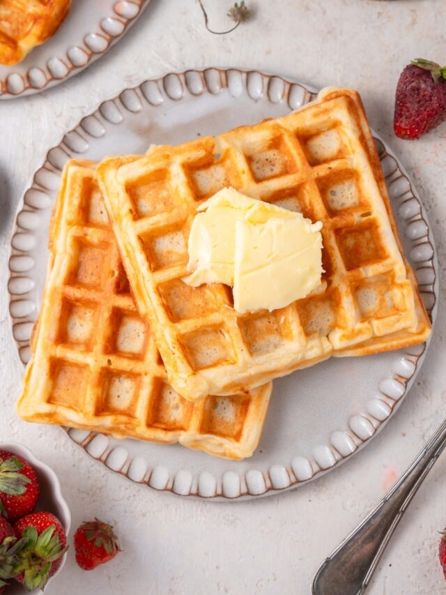 Two cottage cheese waffles on a ceramic plate topped with butter. The plate is on a marble counter with another plate of waffles and a plate of strawberries off to the side.