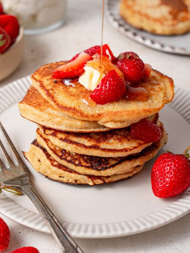 Syrup being poured over a stack of cottage cheese pancakes topped with butter and strawberries.