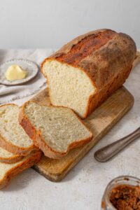 A cottage cheese bread loaf, partially cut into slices and served over a wooden board with a plate of butter in the back.