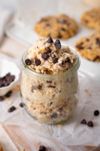 Cottage cheese chocolate chip cookie dough in a glass pot, in front of a tray of chocolate chip cookies.