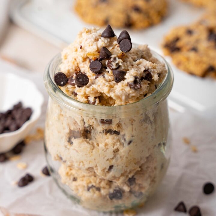 Cottage cheese chocolate chip cookie dough in a glass pot, in front of a tray of chocolate chip cookies.
