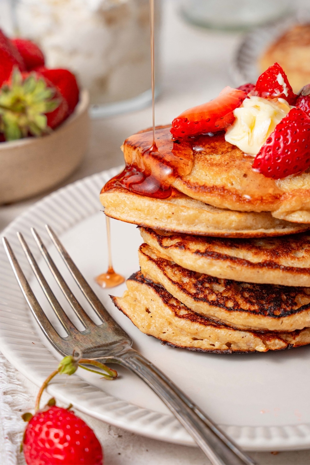 Syrup being poured over and dripping from a stack of five cottage cheese pancakes garnished with butter and strawberries.