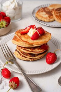 Five cottage cheese pancakes on a plate, garnished with butter and strawberries and served with a fork.