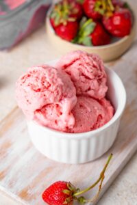 Three scoop of strawberry cottage cheese ice cream in a ramekin next to a bowl of fresh strawberries.