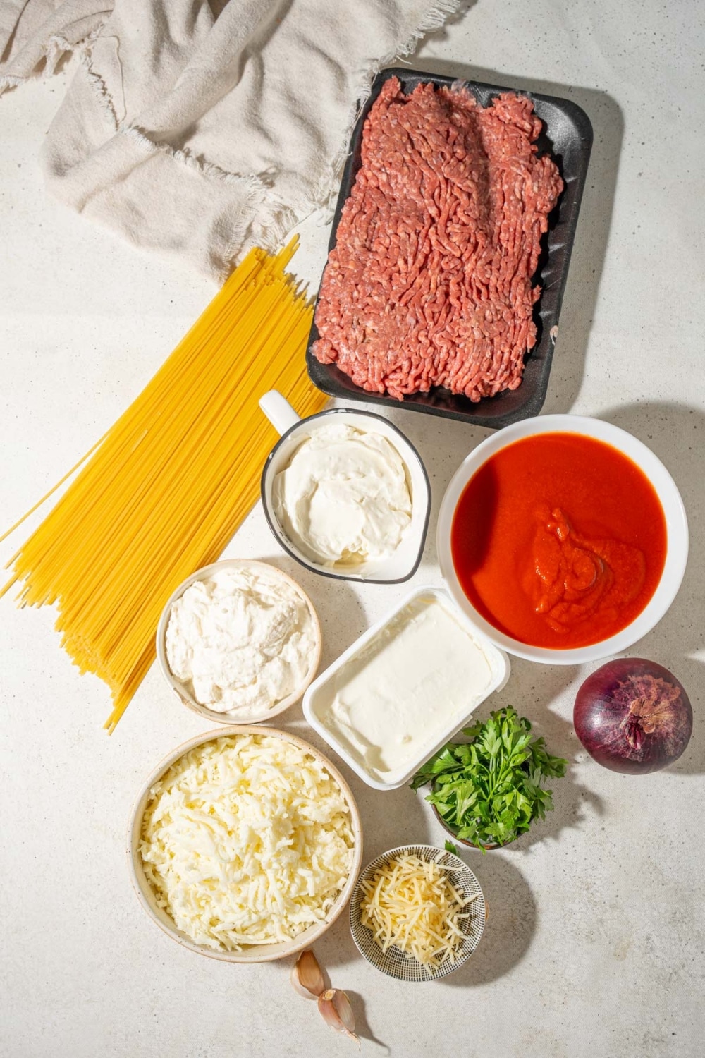 A package of ground beef, bowls of marinara sauce, cottage cheese, sour cream, shredded mozzarella cheese, shredded parmesan, cream cheese, and parsley, a red onion, and dried spaghetti.