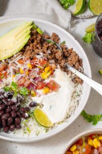 A taco bowl with meat, veggies, beans, rice, a slice of lime, and cottage cheese sauce with a spoon.
