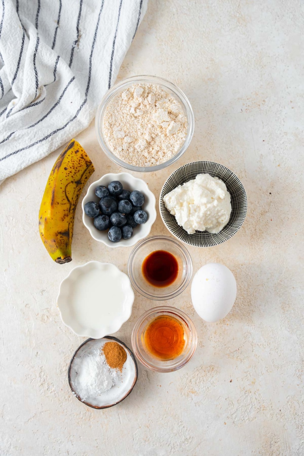 Bowls of oat flour, fresh blueberries, cottage cheese, vanilla extract, almond milk, maple syrup, baking powder, and cinnamon, a banana, and an egg.