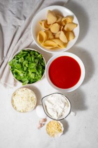 Bowls of cooked pasta shells, marinara sauce, spinach, shredded mozzarella, cottage cheese, and parmesan cheese, an egg, and two garlic cloves.