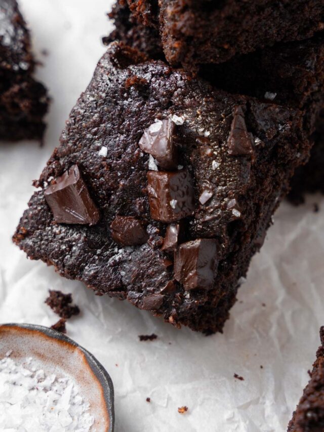 A cottage cheese brownie topped with flaky sea salt, laying on a stack of more cottage cheese brownies next to a small bowl of flaky sea salt.