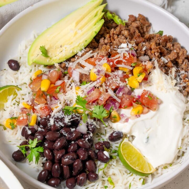 A taco bowl with taco meat, sliced avocado, rice, veggies, beans, and cottage cheese sauce.