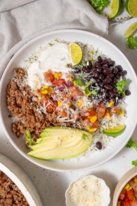 Top view of a taco bowl with cilantro lime rice, black beans, taco meat, corn salsa, sliced avocado, lime, and cottage cheese sauce.