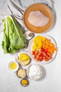 Plates of chicken breast, sliced yellow and orange bell peppers, sliced cherry tomatoes, sliced lemon, bowls of salt, black pepper, garlic powder, cottage cheese, shredded cheese, mustard, and lemon juice, and a head of romaine lettuce.