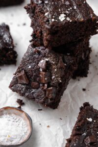 A stack of cottage cheese brownies on parchment paper next to a small bowl of flaky sea salt.