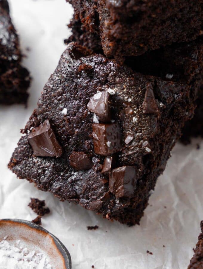A cottage cheese brownie topped with flaky sea salt, laying on a stack of more cottage cheese brownies next to a small bowl of flaky sea salt.