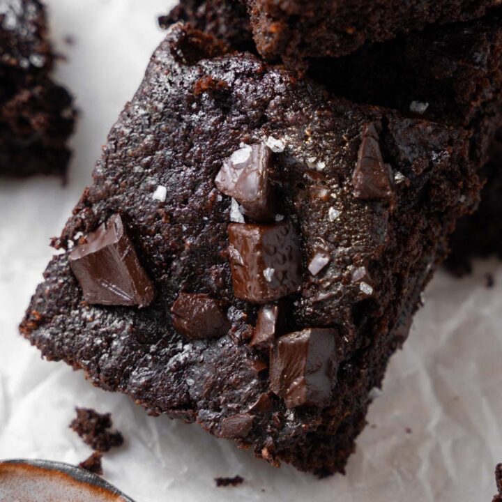 A cottage cheese brownie topped with flaky sea salt, laying on a stack of more cottage cheese brownies next to a small bowl of flaky sea salt.