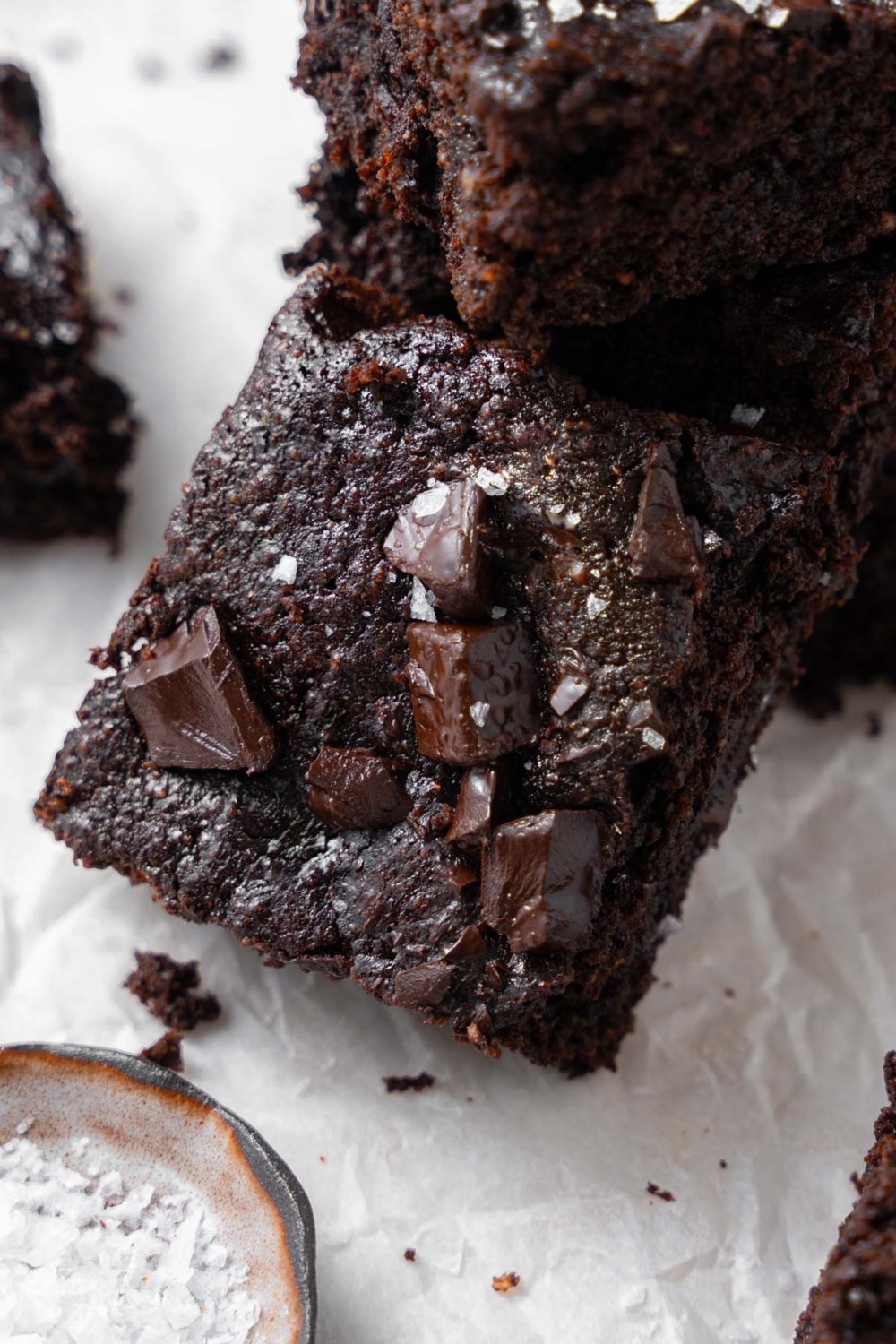 A cottage cheese brownie topped with flaky sea salt, laying on a stack of more cottage cheese brownies next to a small bowl of flaky sea salt.