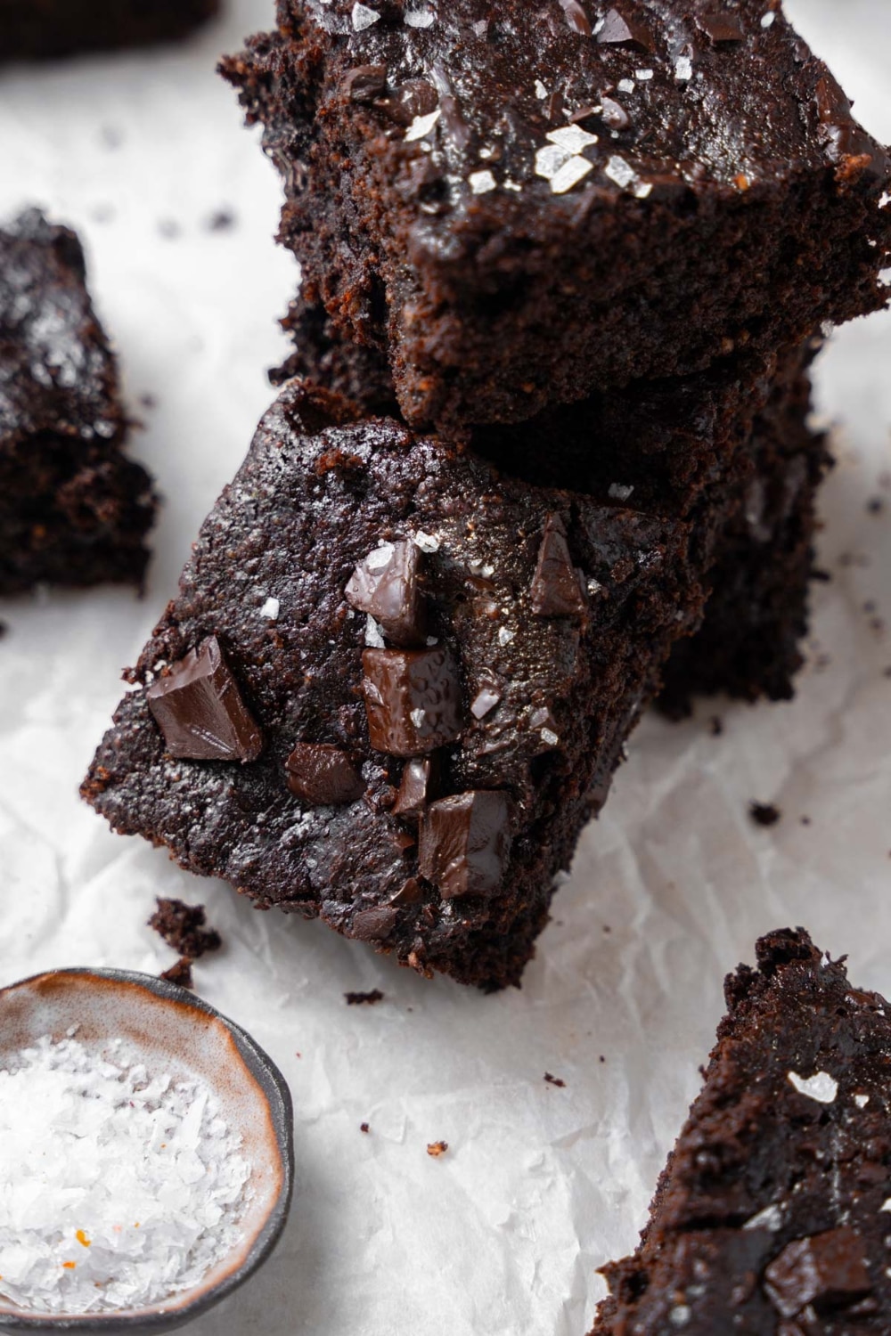 A stack of cottage cheese brownies on parchment paper next to a small bowl of flaky sea salt.