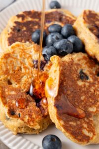Maple syrup being drizzled on cottage cheese banana blueberry pancakes.