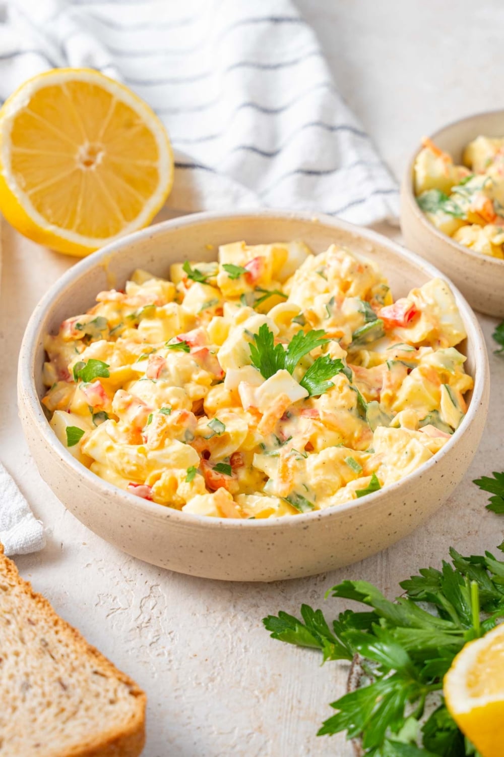 A bowl of cottage cheese egg salad garnished with fresh parsley and served alongside a sliced lemon, a slice of bread, and another bowl of egg salad.