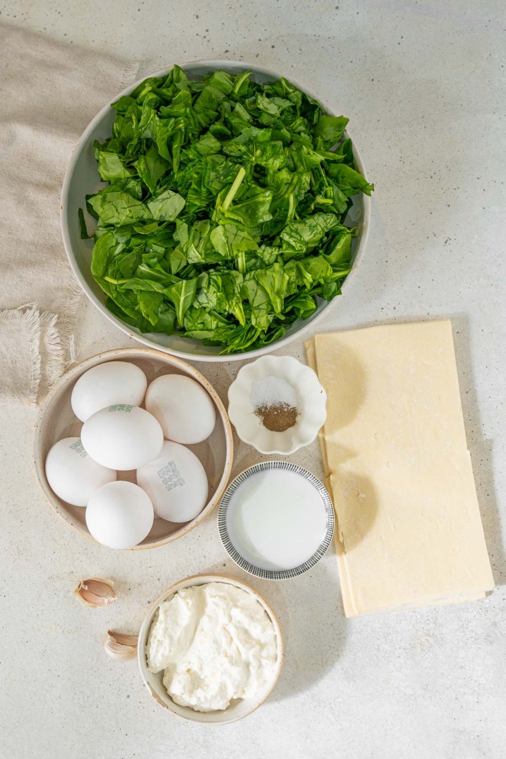 Bowls of fresh spinach, eggs, cottage chees, whole milk, salt, and pepper, puff pastry sheets, and garlic cloves.