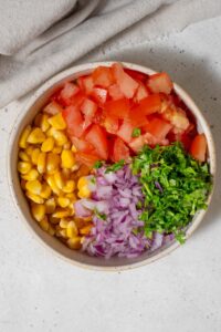 A bowl of corn kernels, minced red onion, chopped cilantro, and diced tomatoes.