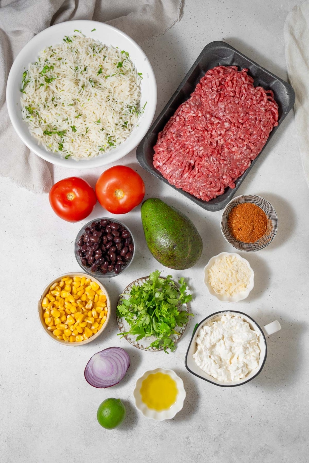 A package of ground beef, a bowl of cilantro lime rice, two tomatoes, an avocado, bowls of taco seasoning, shredded cheese, black beans, corn, cilantro, cottage cheese, and lemon juice, a sliced red onion, and a lime.